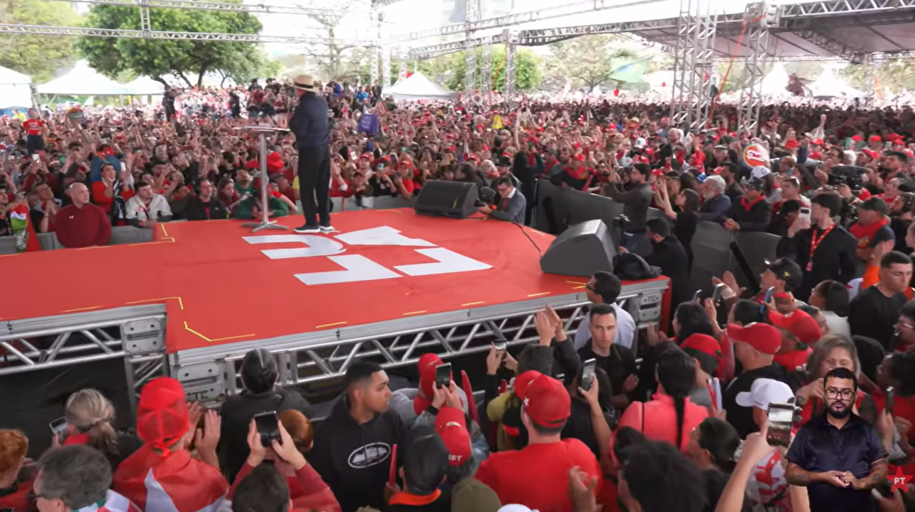 Imagem de um grande evento ao ar livre, com uma pessoa em pé sobre um palco vermelho diante de uma multidão numerosa. Muitas pessoas estão com os braços levantados, aplaudindo e segurando celulares para registrar o momento. Ao redor do palco há estruturas metálicas, equipamentos de som e tendas.
