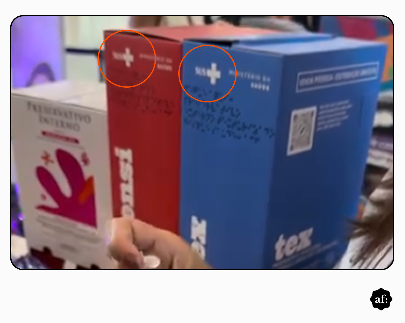 Três caixas de papelão estão dispostas sobre uma superfície, atuando como dispensadores de insumos. À esquerda, uma caixa predominantemente branca com detalhes em rosa e laranja exibe o texto ‘PRESERVATIVO INTERNO’. Ao centro e à direita, encontram-se duas caixas mais altas, uma vermelha e uma azul, ambas contendo o logotipo do SUS com uma cruz branca e a inscrição ‘MINISTÉRIO DA SAÚDE’. A caixa azul apresenta um código QR, o texto ‘VENDA PROIBIDA - DISTRIBUIÇÃO GRATUITA’, as letras ‘tex’ na parte inferior. Em primeiro plano, na parte inferior, uma mão com um anel prateado segura um pequeno objeto transparente. No limite do canto inferior direito, é visível parte do cabelo escuro e da camiseta branca com letras pretas de uma pessoa, enquanto outras figuras aparecem desfocadas ao fundo.