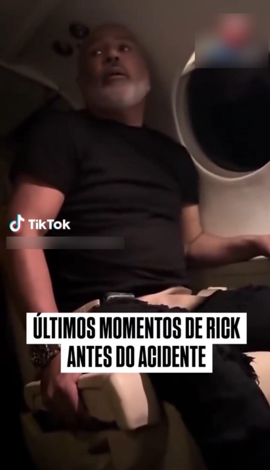A imagem mostra um homem de pele parda, calvo, com barba e bigode grisalhos, vestindo uma camiseta preta de mangas curtas e calça preta com detalhes rasgados no joelho. Ele está sentado dentro da cabine de uma aeronave executiva ou jatinho, segurando o braço da poltrona com a mão direita. Do lado esquerdo da imagem, há o logotipo da rede social TikTok. Na parte inferior, sobrepostas à cena, aparecem duas faixas brancas com o texto em caixa alta: ‘ÚLTIMOS MOMENTOS DE RICK ANTES DO ACIDENTE’.