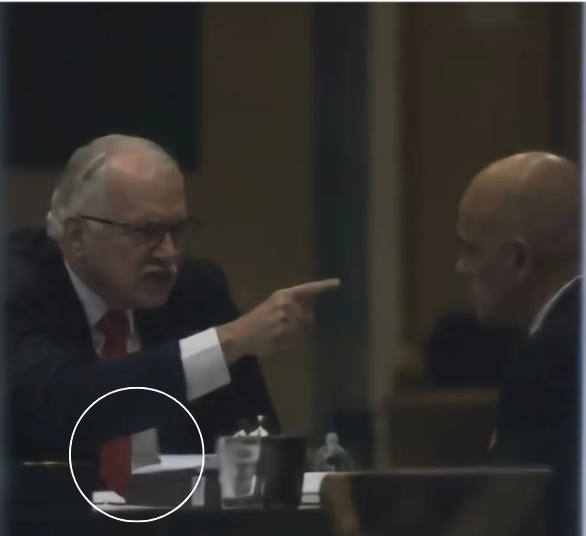 Dois homens — identificados como os ministros do STF Edson Fachin e Alexandre de Moraes —, vestindo ternos escuros e camisas brancas, estão sentados em uma mesa em um ambiente interno. Fachin, homem de pele clara com cabelos grisalhos e óculos, está à esquerda e aponta o dedo indicador para Moraes, um homem de pele clara e calvo, que está à direita e olha para ele. A expressão facial de Fachin sugere tensão. No primeiro plano da imagem, há uma mesa com papéis, copos e outros objetos desfocados. Um círculo branco destaca parte da área inferior esquerda da imagem. A qualidade da imagem é granulada e ligeiramente desfocada.