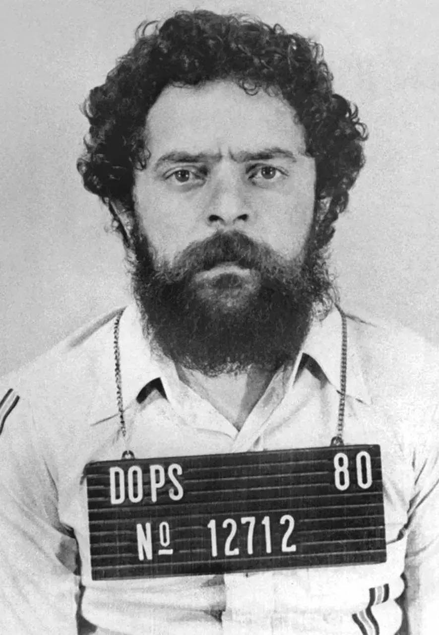 Fotografia em preto e branco mostra Lula da Silva de cabelos e barba escuros, olhando diretamente para a câmera com expressão séria. Ele veste uma camisa clara e tem uma placa de identificação policial pendurada no pescoço com as inscrições ‘DOPS, 80, Nº 12712’.