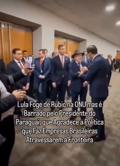 O presidente Luiz Inácio Lula da Silva, está de pé em um corredor ao lado do presidente do Paraguai, Santiago Peña — homem de pele clara, cabelos escuros penteados para trás e terno escuro com gravata vermelha, e de Beto Ovelar, homem de pele clara, cabelos escuros e terno escuro, visto de costas enquanto cumprimenta Lula. Na parte inferior da imagem há um texto sobreposto em letras brancas que diz: ‘Lula Foge de Rubio na ONU mas é Barrado pelo Presidente do Paraguai, que Agradece a Política que Faz Empresas Brasileiras Atravessarem a Fronteira’.