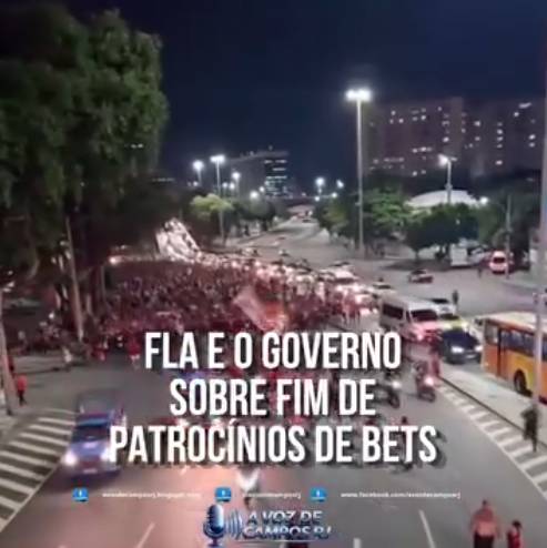 Uma captura de tela exibe uma via urbana noturna iluminada por postes de luz, com árvores à esquerda e edifícios ao fundo. Na metade esquerda da pista e na calçada adjacente, concentra-se uma aglomeração de pessoas que vestem predominantemente roupas vermelhas; devido à distância e à resolução da imagem, não é possível identificar características físicas individuais. Na metade direita da via, observa-se o tráfego de veículos, incluindo carros, uma van branca e um ônibus amarelo. Sobreposto à região central da imagem, há o texto em letras maiúsculas brancas 'FLA E O GOVERNO SOBRE FIM DE PATROCÍNIOS DE BETS'. Na base da composição, visualizam-se ícones de redes sociais alinhados a um logotipo de microfone azul acompanhado da inscrição 'A VOZ DE CAMPOS RJ'. 