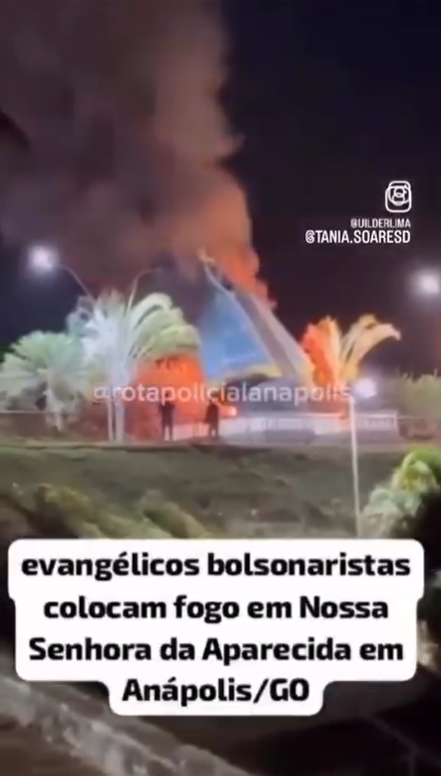 Imagem noturna mostra um monumento em chamas, cercado por iluminação pública e palmeiras, com uma grande nuvem de fumaça subindo ao céu escuro. Na base da estrutura, duas silhuetas escuras de pessoas aparecem em pé, sem características físicas ou vestimentas distinguíveis devido à distância e à iluminação. Uma caixa de texto branca na parte inferior com a frase escrita em letras pretas: 'evangélicos bolsonaristas colocam fogo em Nossa Senhora da Aparecida em Anápolis/GO'. 