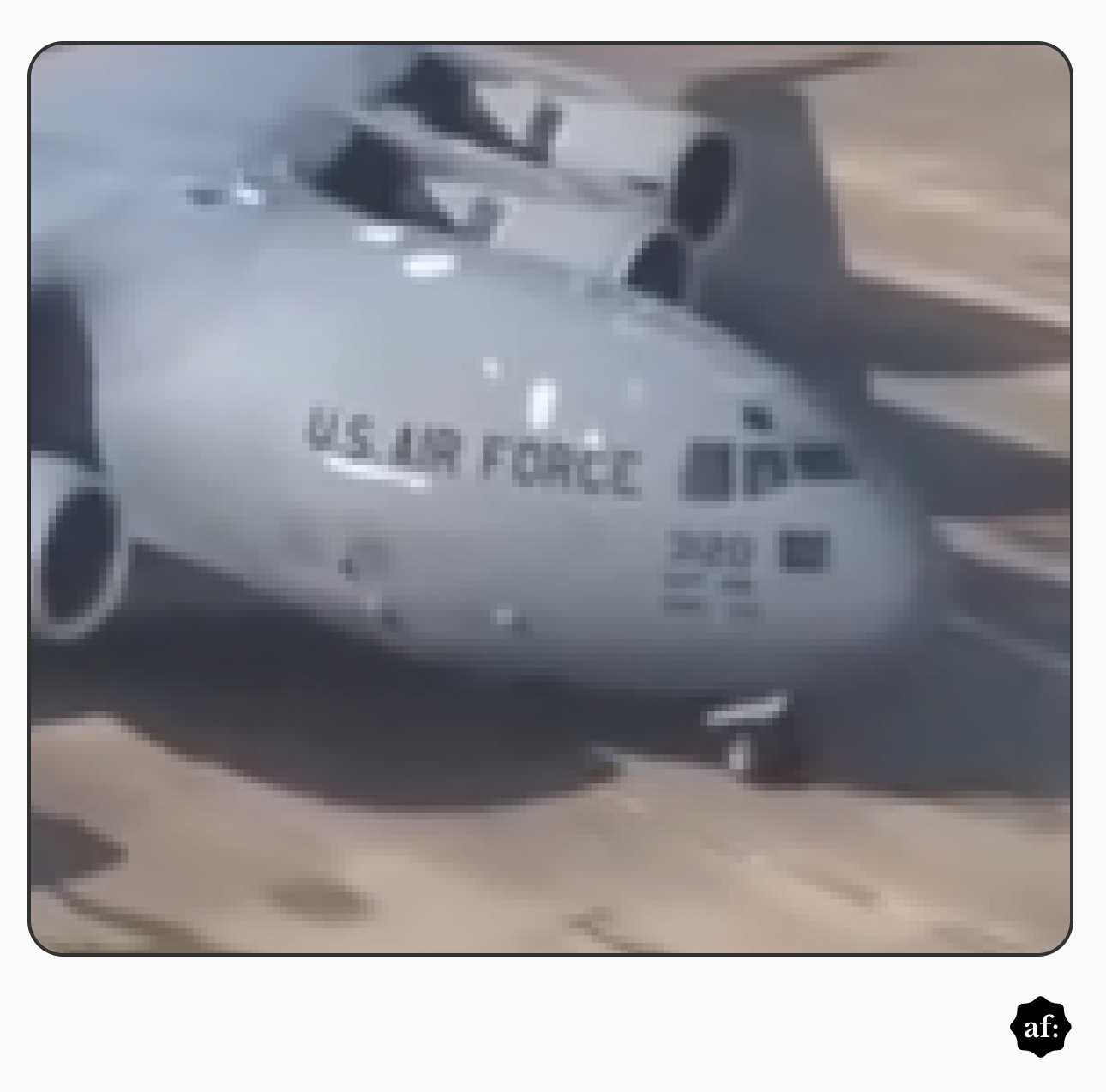Uma fotografia de baixa resolução exibe em primeiro plano a seção frontal de uma grande aeronave de cor cinza-claro em solo. Na lateral da fuselagem, próximo às janelas da cabine de pilotagem, encontra-se o texto 'U.S. AIR FORCE' impresso em letras maiúsculas escuras, acompanhado de perto por números parcialmente legíveis. Na parte superior do avião, são visíveis grandes estruturas cilíndricas associadas aos motores, e, na região inferior, observa-se o pneu do trem de pouso dianteiro apoiado em uma pista plana de tom terroso. No limite do canto inferior direito da composição, sobre uma margem branca externa à fotografia, há um pequeno logotipo circular preto contendo as letras brancas 'af:'.