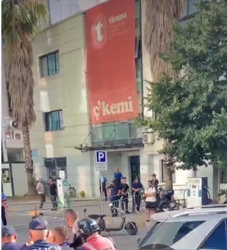 A imagem mostra a fachada de um prédio claro com janelas de vidros escuros e uma palmeira no lado esquerdo. Na parede frontal do edifício, há um painel vertical de cor Laranja onde se lê no topo a palavra 'Tirana' ao lado do logótipo com a letra 't' dentro de círculos, seguido pelo texto 'European Youth Capital 22', e na parte inferior a palavra 'ç'kemi' em letras grandes.