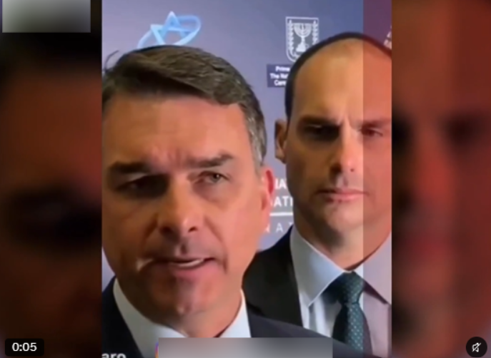 Print de vídeo de Flávio Bolsonaro e Eduardo Bolsonaro na frente de um fundo azul. Ambos estão de terno. Eduardo olha na direção de Flávio, que fala em direção à camera.