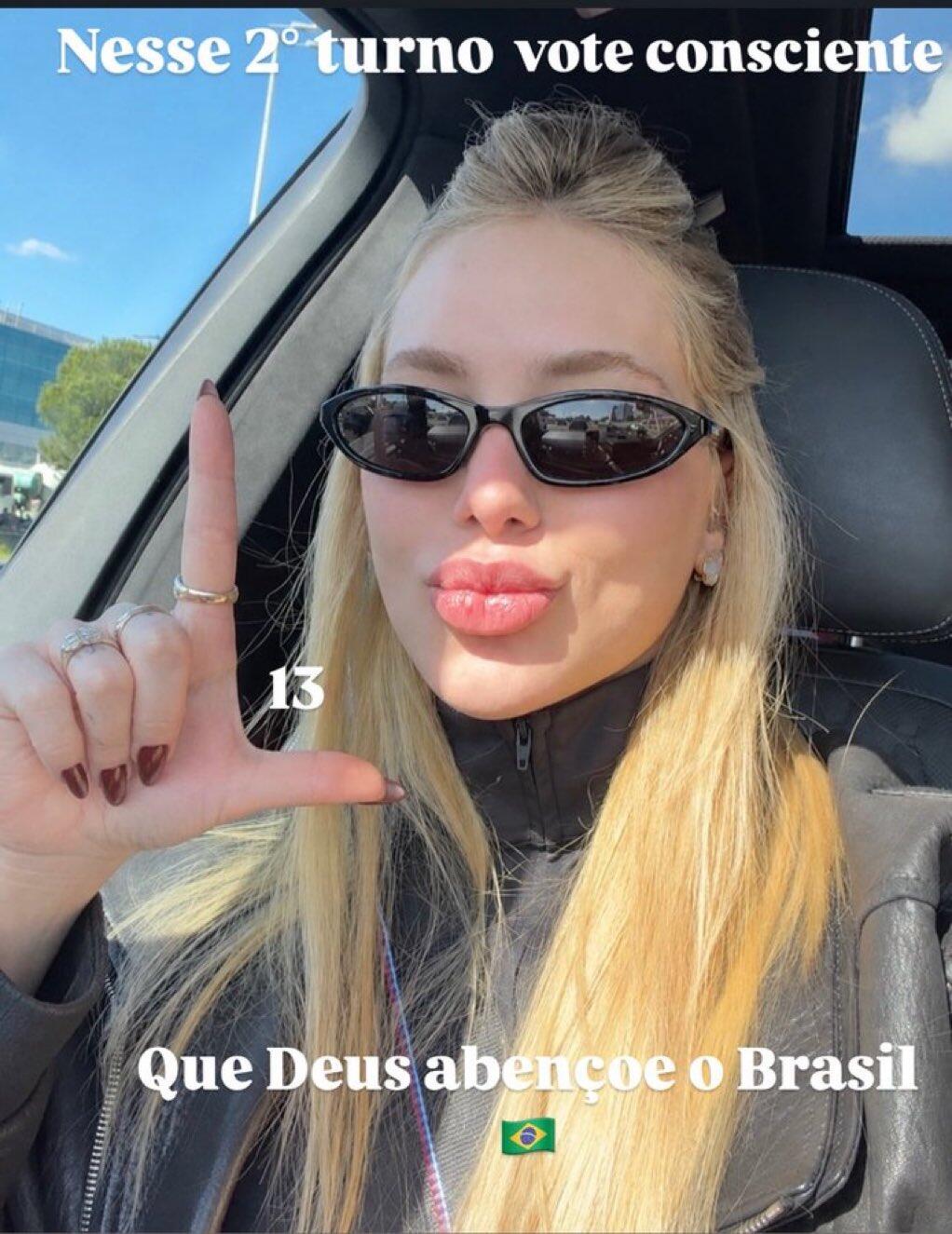 A imagem exibe uma selfie de uma mulher de pele clara, lábios volumosos e longos cabelos loiros, vestindo uma jaqueta preta de gola alta e óculos de sol escuros ovais, sentada no interior de um veículo. Ela faz um gesto com a mão direita, estendendo o dedo indicador para cima e o polegar para o lado, formando a letra 'L', e exibe unhas escuras e anéis. Sobre a fotografia, há textos brancos inseridos: na parte superior, lê-se 'Nesse 2º turno vote consciente'; ao lado da mão, o número '13'; e na base, a frase 'Que Deus abençoe o Brasil' seguida por um pequeno emoji da bandeira do país.