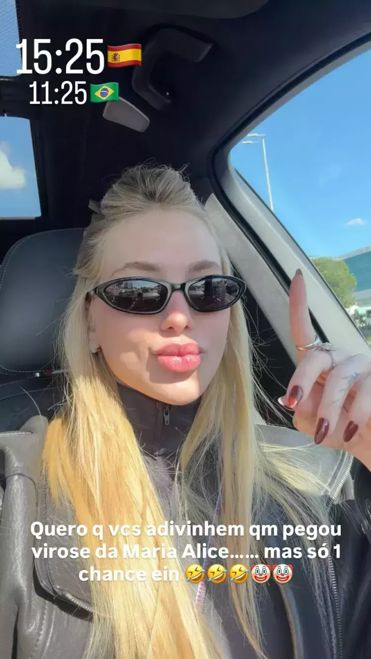 A imagem exibe uma selfie de uma mulher de pele clara, lábios volumosos e longos cabelos loiros, vestindo uma jaqueta preta de gola alta e óculos de sol escuros ovais, sentada no interior de um veículo. Com a mão direita, que apresenta unhas escuras e anéis, ela mantém o dedo indicador apontado para cima. No canto superior esquerdo da fotografia, estão inseridos os textos '15:25' ao lado de um emoji da bandeira da Espanha e '11:25' com um emoji da bandeira do Brasil. Na parte inferior, há uma frase sobreposta em letras brancas que diz: 'Quero q vcs adivinhem qm pegou virose da Maria Alice...... mas só 1 chance ein', seguida por quatro emojis de rostos rindo e um emoji de palhaço