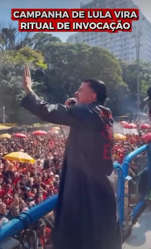 A imagem mostra uma pessoa de pele clara e cabelo escuro curto, vestindo um longo casaco preto com detalhes em vermelho nas costas — que incluem letras parciais e o desenho de um tridente —, posicionada em uma estrutura elevada delimitada por barras azuis. Essa pessoa está com o braço esquerdo erguido e a mão espalmada, de frente para uma grande multidão aglomerada na rua abaixo, e parece segurar um microfone próximo ao rosto com a outra mão. À sua direita, no mesmo nível elevado, há um homem de pele clara, com barba, usando um chapéu preto e camiseta listrada, que segura um aparelho celular apontado para a frente. Ao fundo, além da aglomeração de pessoas que portam alguns guarda-sóis coloridos, observam-se copas de árvores e altos edifícios sob a luz do dia. Na parte superior da imagem, há um texto destacado em letras brancas sobre fundo vermelho onde se lê: 'CAMPANHA DE LULA VIRA RITUAL DE INVOCAÇÃO'.