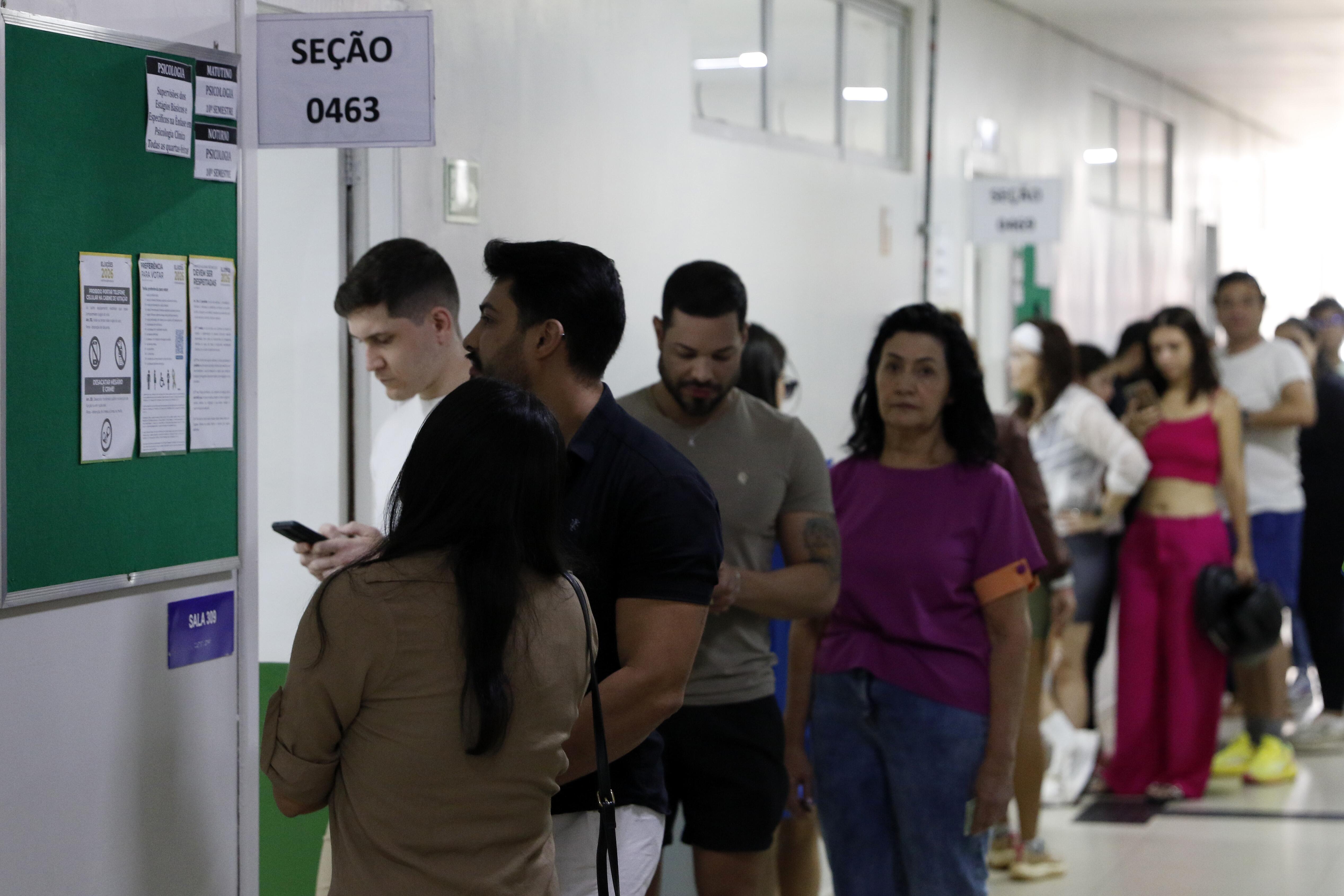 A imagem mostra uma fila de pessoas em um corredor de paredes brancas, com portas e janelas de vidro na parte superior. À esquerda, há uma placa com a inscrição 'SEÇÃO 0463' acima da entrada de uma sala. Ao lado, um mural verde apresenta avisos e orientações sobre as eleições de 2026. Em primeiro plano, uma mulher de cabelos longos e escuros, vestindo uma blusa marrom, aparece de costas. À sua frente, um homem de camiseta preta e calça branca está de perfil. Atrás dele, um homem de camiseta branca olha para um celular. Na sequência da fila, aparecem um homem de camiseta cinza e bermuda preta, uma mulher de blusa roxa e calça jeans e outras pessoas. Ao fundo, uma mulher veste roupas cor-de-rosa e segura um objeto preto. O corredor tem piso claro e iluminação branca.