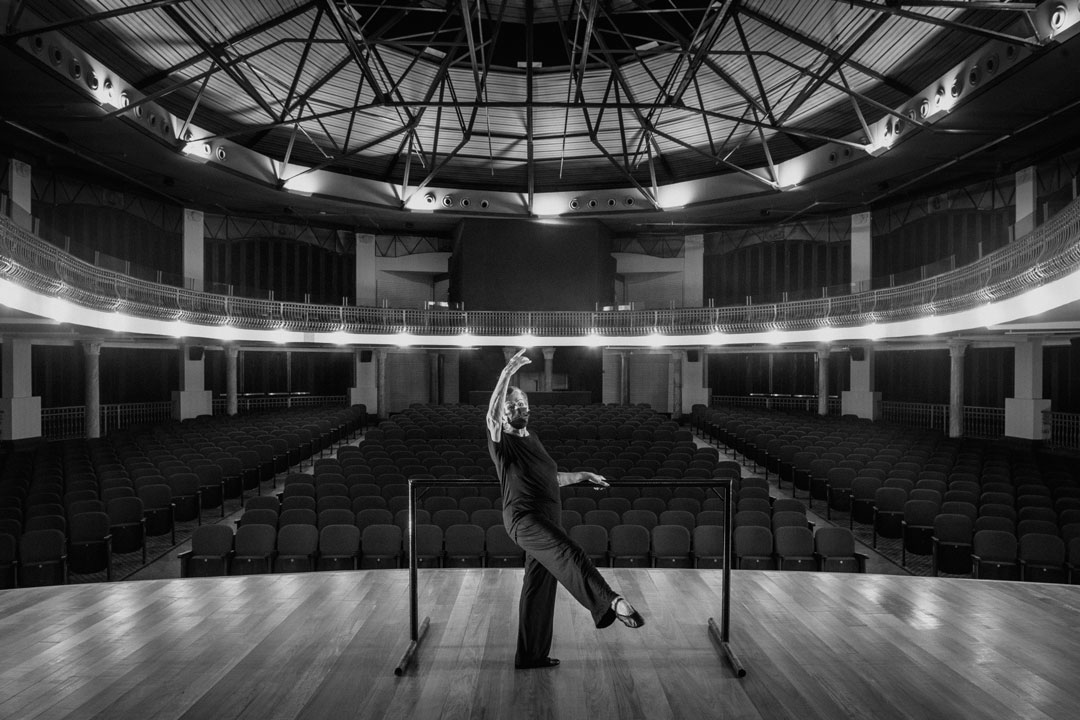 De máscara, uma senhora treina balé em um teatro vazio.
