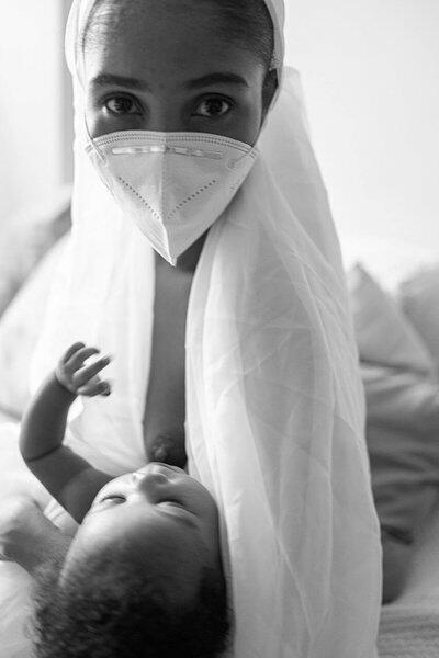 De máscara, mulher amamenta o bebê nos seus braços.